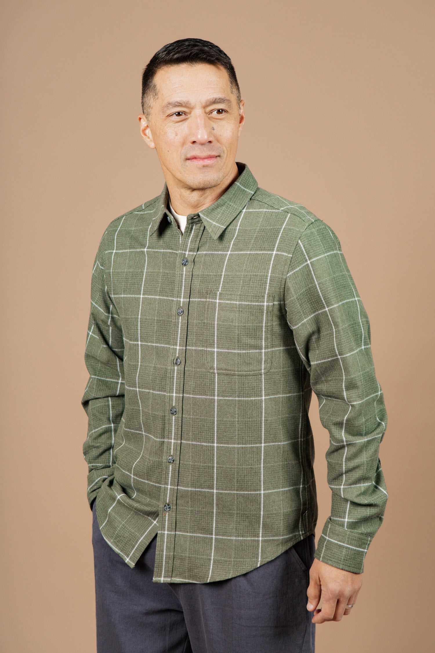 A man in the Bridge & Burn Larson Shirt in Rosemary Twill Windowpane and gray pants stands against a beige background, looking slightly left with a neutral expression.