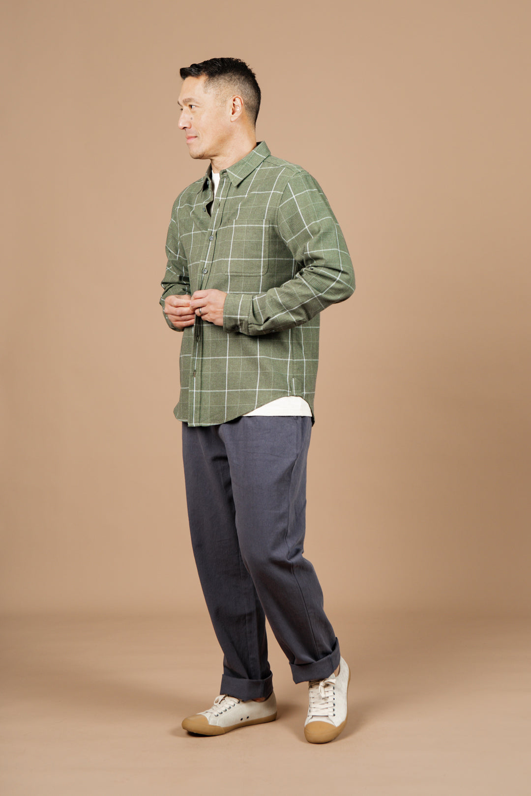 A man stands smiling in a studio with a beige background, wearing Bridge & Burn's Larson Shirt in Rosemary Twill Windowpane over a white tee, loose blue-gray pants, and classic white sneakers with tan soles. He looks to his left.