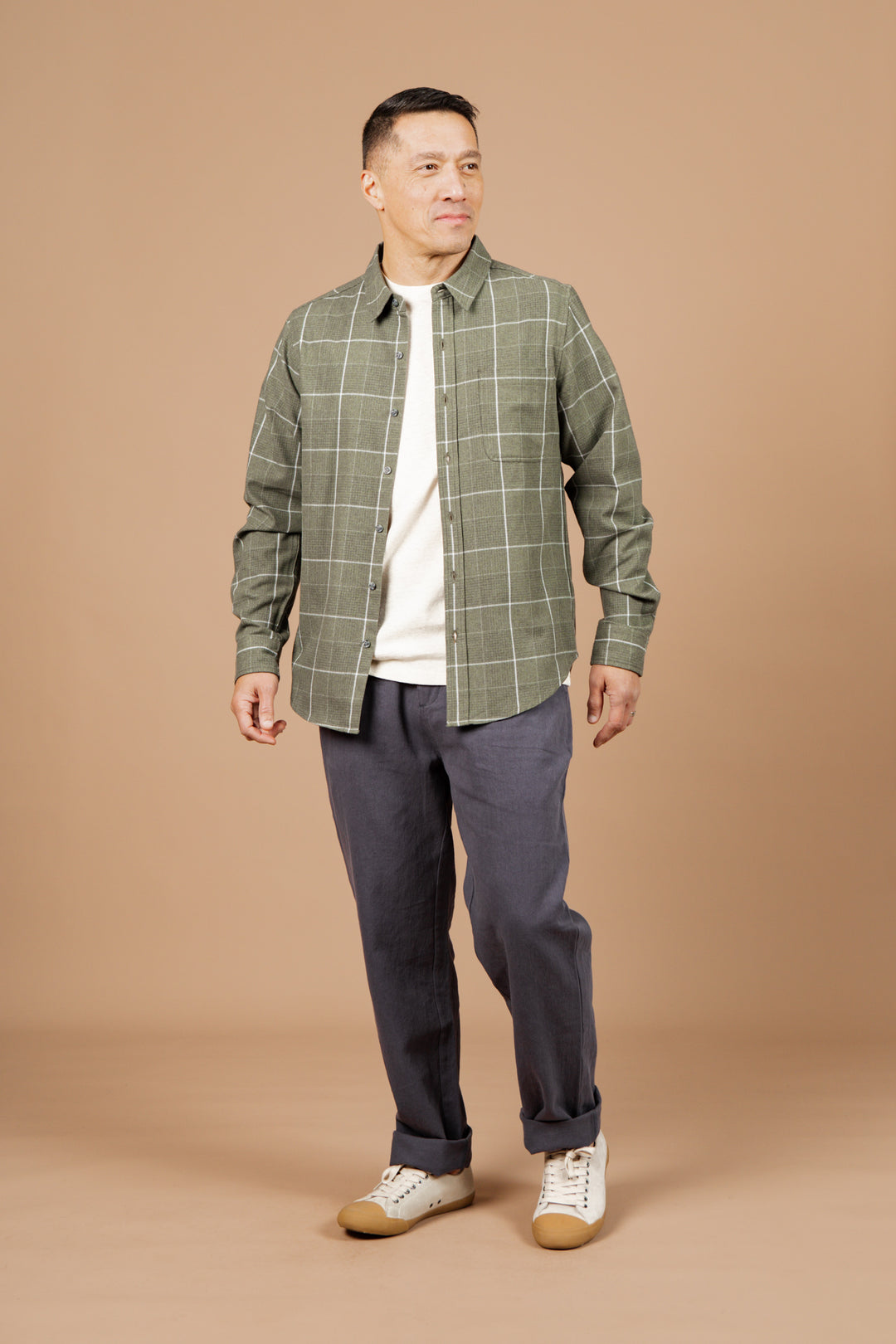 A man stands against a beige background wearing the Bridge & Burn Larson Shirt in Rosemary Twill Windowpane over a white t-shirt, with gray pants and white sneakers. He looks to the side with a slight smile.