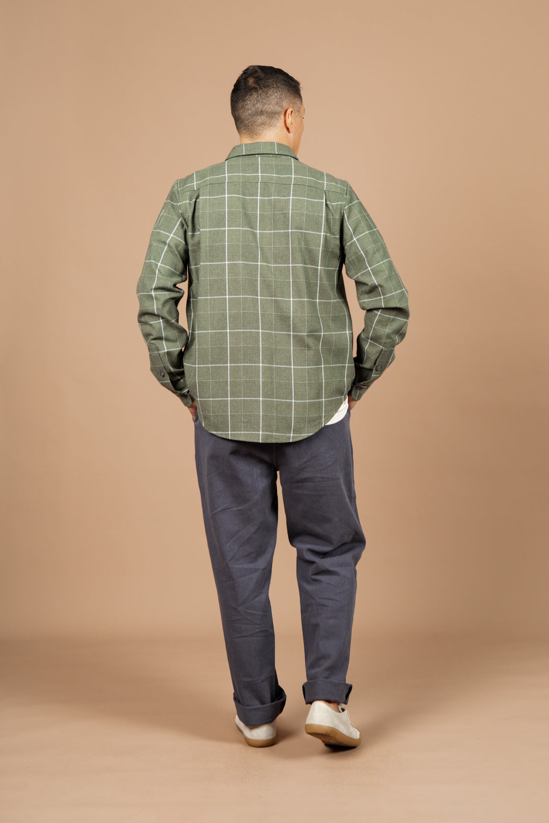 A person faces away from the camera wearing the Bridge & Burn Larson Shirt in Rosemary Twill Windowpane, paired with gray pants and white shoes, set against a plain beige background.