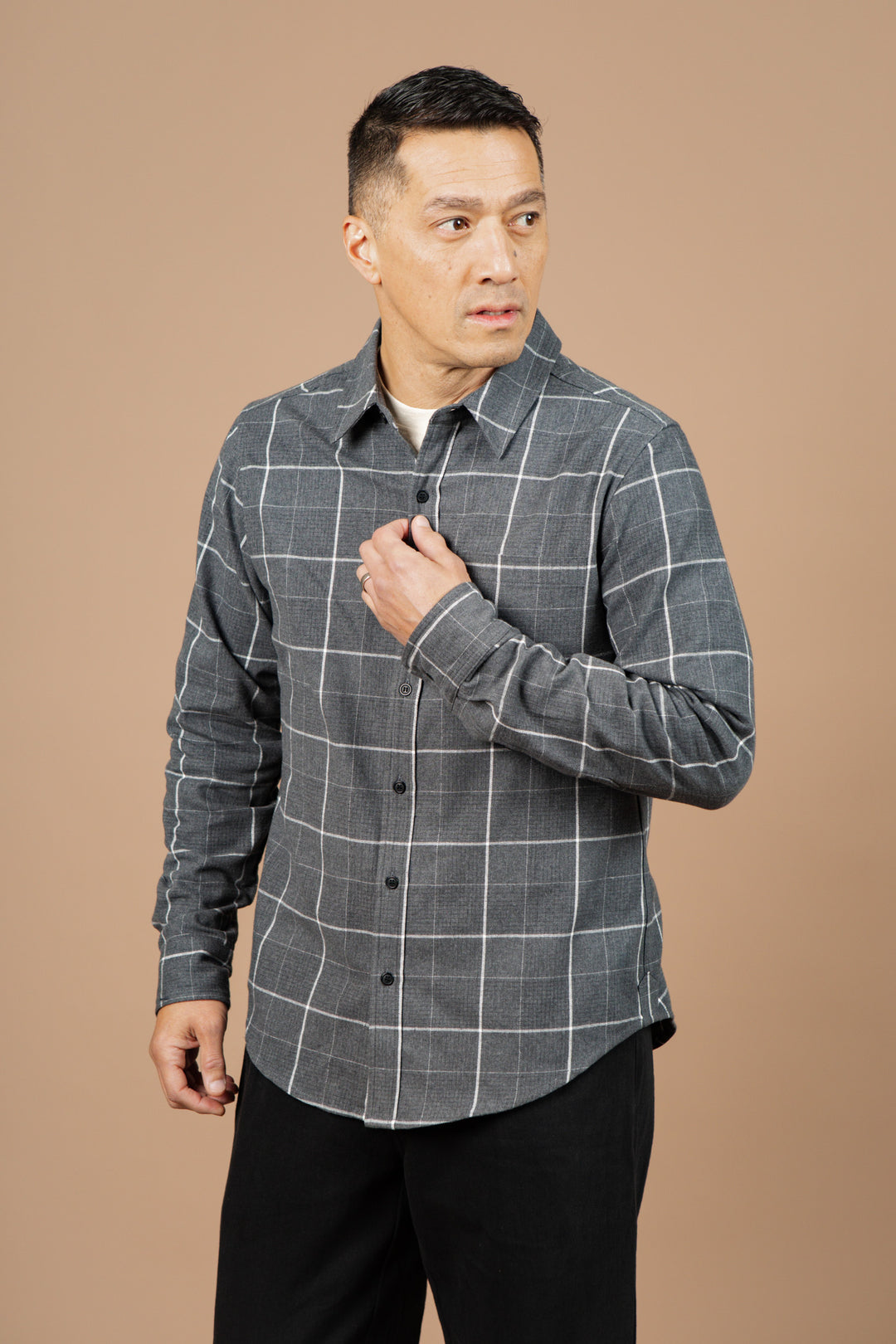 A man wearing the Bridge & Burn Larson Shirt in Shadow Twill Windowpane and black pants stands against a plain beige background, looking to his right while adjusting his shirt with one hand.