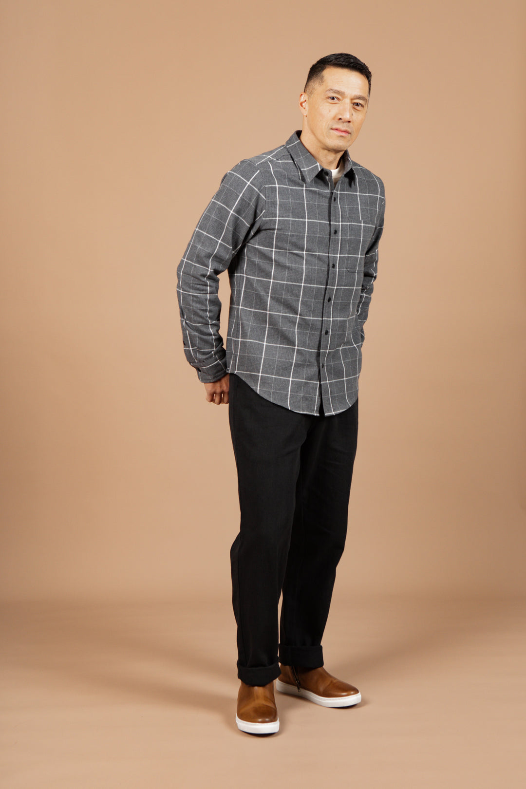 A man with short dark hair stands on a tan background, wearing the Bridge & Burn Larson Shirt in Shadow Twill Windowpane, paired with black pants and brown shoes. He looks slightly to the side with a neutral expression.
