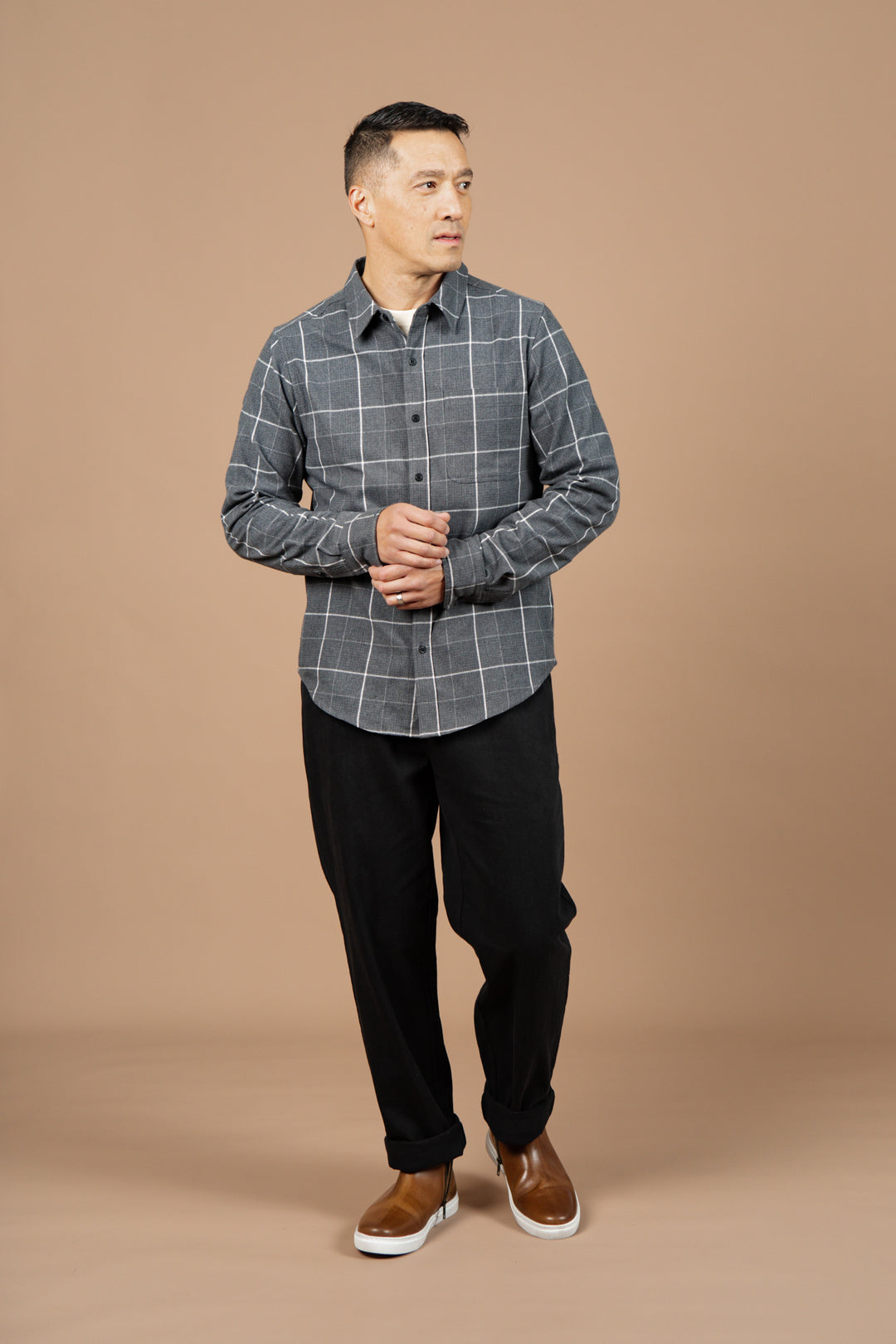 A man stands against a beige background wearing the Bridge & Burn Larson Shirt in Shadow Twill Windowpane, paired with black pants and brown slip-ons. He looks slightly left with his hands loosely clasped.