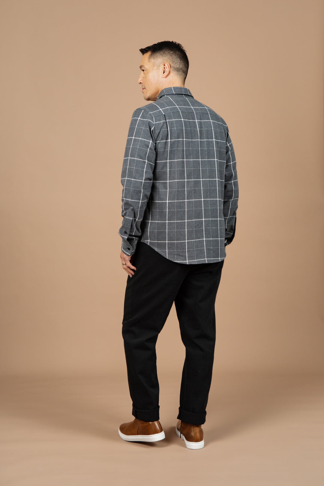 A man stands facing away, wearing the Bridge & Burn Larson Shirt in Shadow Twill Windowpane, black pants, and brown sneakers against a plain beige background.