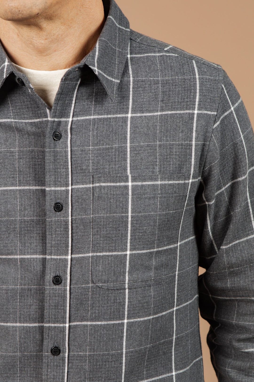 A close-up of a person in the Bridge & Burn Larson Shirt in Shadow Twill Windowpane, a classic fit gray brushed flannel button-up with white checks, layered over a light undershirt against a beige background. Face partially visible.