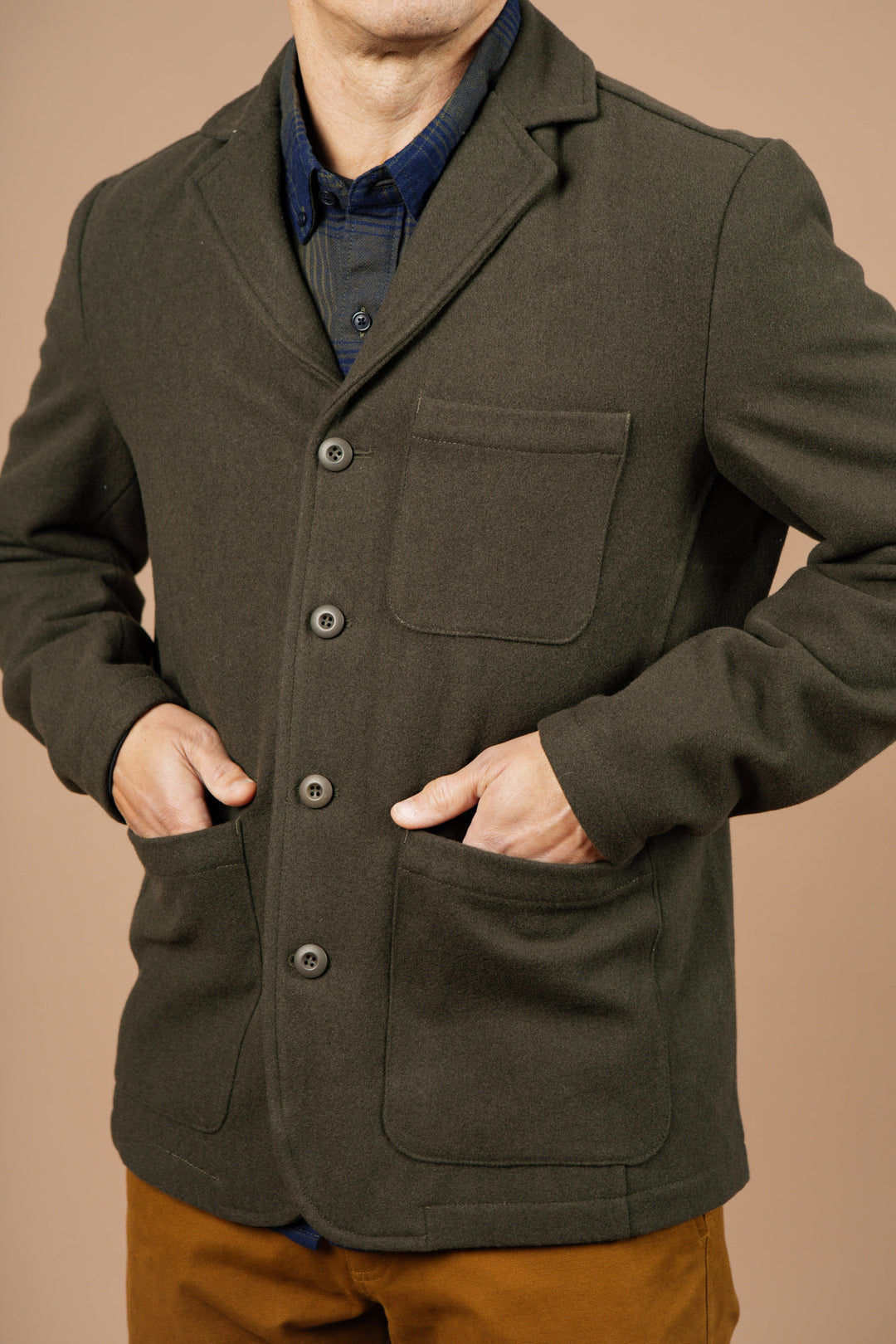 A person wearing the Bridge & Burn Nate Chore Blazer in Basque Green Wool over a dark shirt and brown pants stands against a beige background with hands in the blazer’s pockets.