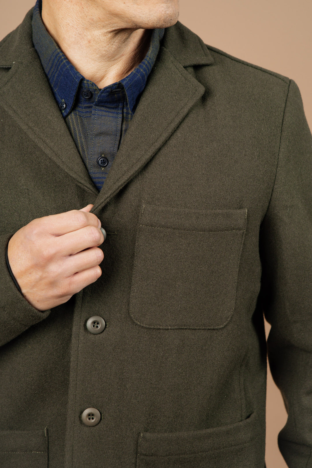 A person wearing the Bridge & Burn Nate Chore Blazer in Basque Green Wool over a dark blue plaid shirt adjusts the lapel with their left hand, their face partially visible and mostly turned away from the camera.