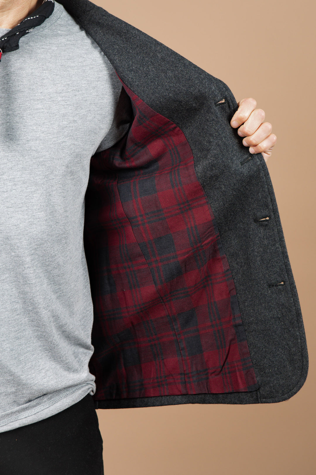 A person holds open the Bridge & Burn Nate Chore Blazer in Onyx Wool, revealing its red and black plaid lining over a light gray shirt with a plain beige background—an ideal standout piece for winter.