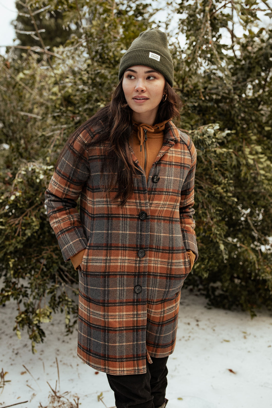 A woman stands outside in the snow, hands in her pockets, wearing the Bridge & Burn Alberta Car Coat in Dorset Brushed Plaid with a green beanie. Snowy trees create a peaceful winter backdrop as she gazes into the distance.