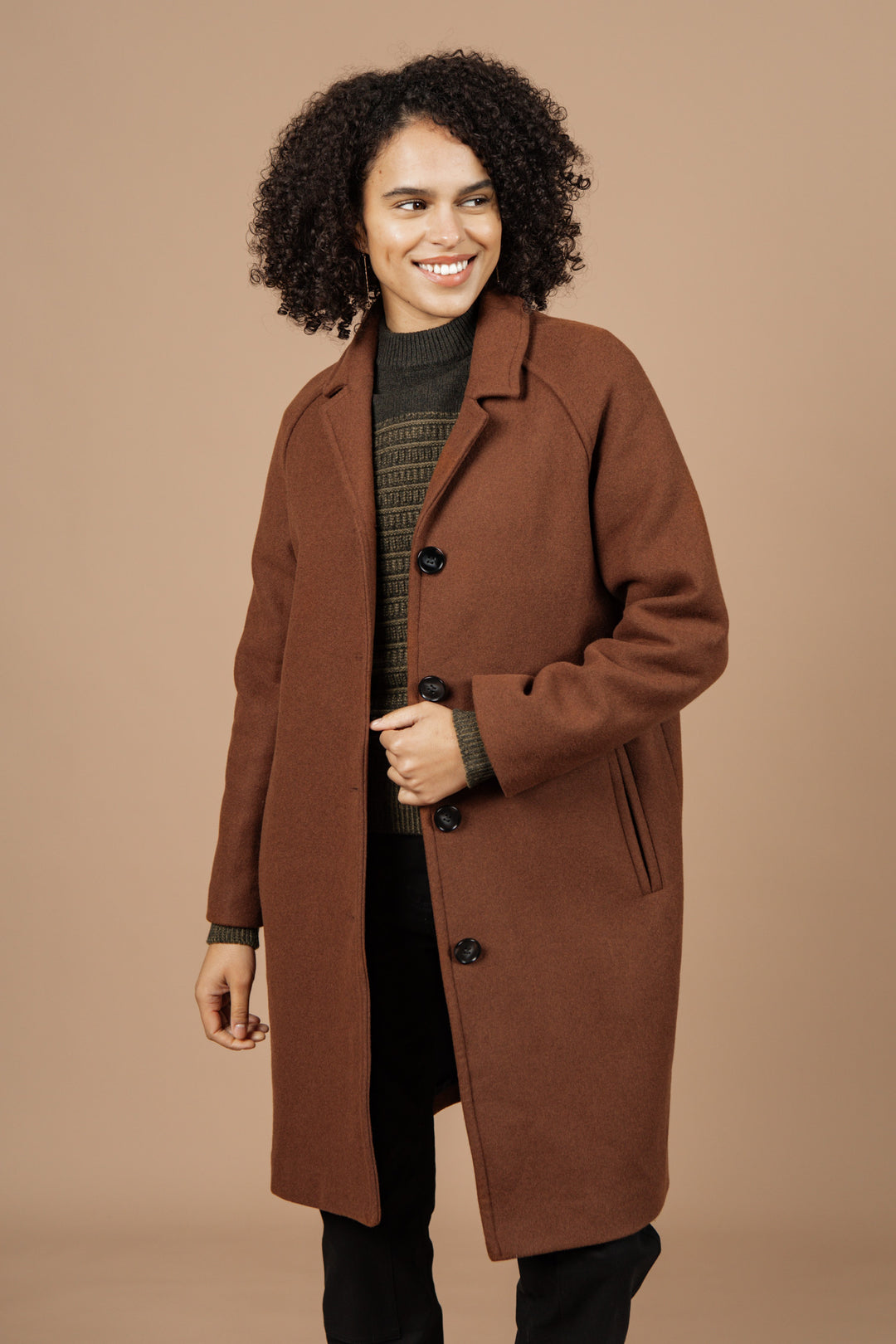 A person with curly hair smiles while wearing the Bridge & Burn Alberta Car Coat in Fired Brick over a green sweater and black pants, standing against a plain light brown background.