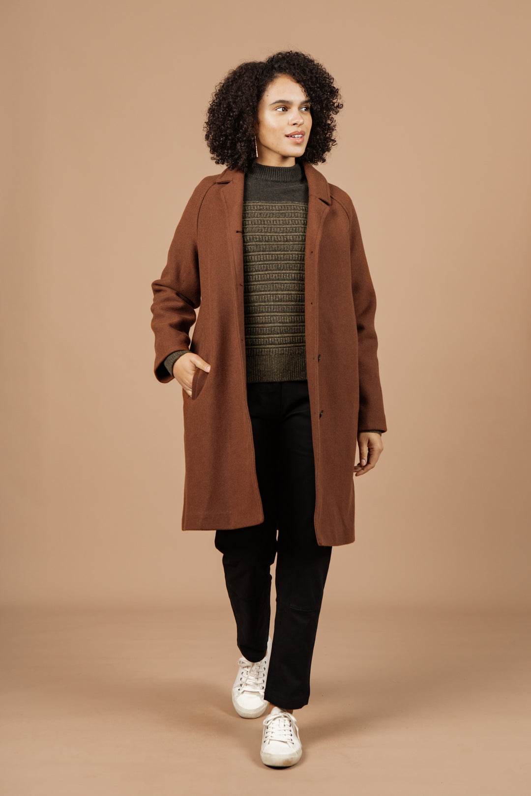 A person with curly hair wears the Bridge & Burn Alberta Car Coat in Fired Brick over a green sweater, black pants, and white sneakers, standing against a plain beige background.