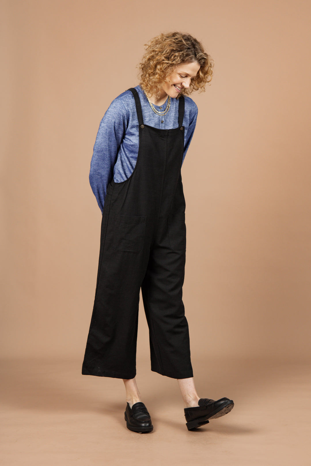 A woman with curly hair stands on a tan background, smiling and looking down with her hands behind her back. She is wearing the Bridge & Burn Edie Overall in Black over a blue long-sleeve shirt and black shoes.