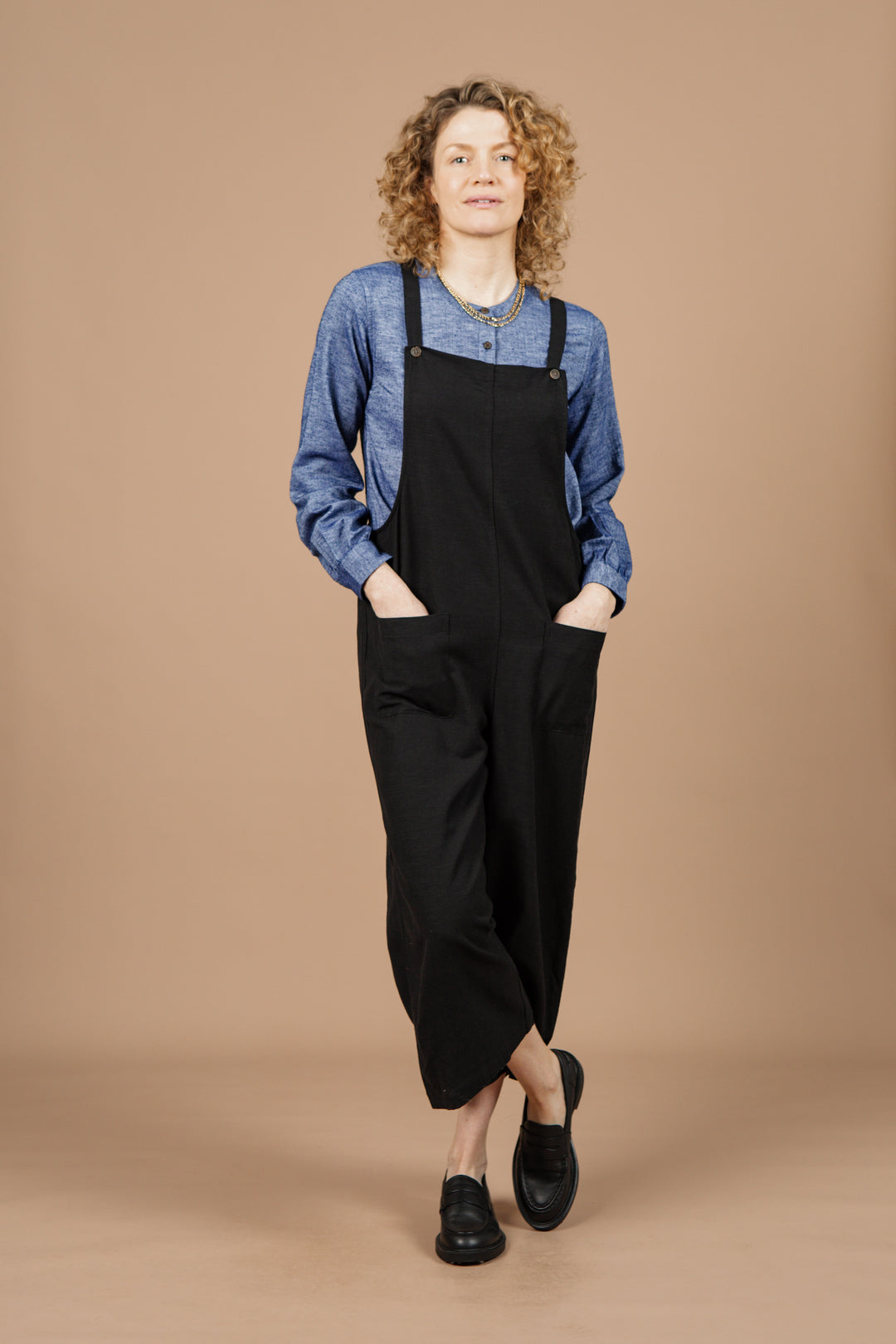 A person with curly hair stands against a beige background, wearing the Bridge & Burn Edie Overall in Black—a pair of wide leg overalls with adjustable straps, crafted from a comfortable linen-cotton blend—styled over a blue long-sleeve shirt and paired with black shoes. Their hands rest in their pockets and one leg is slightly crossed in front of the other.