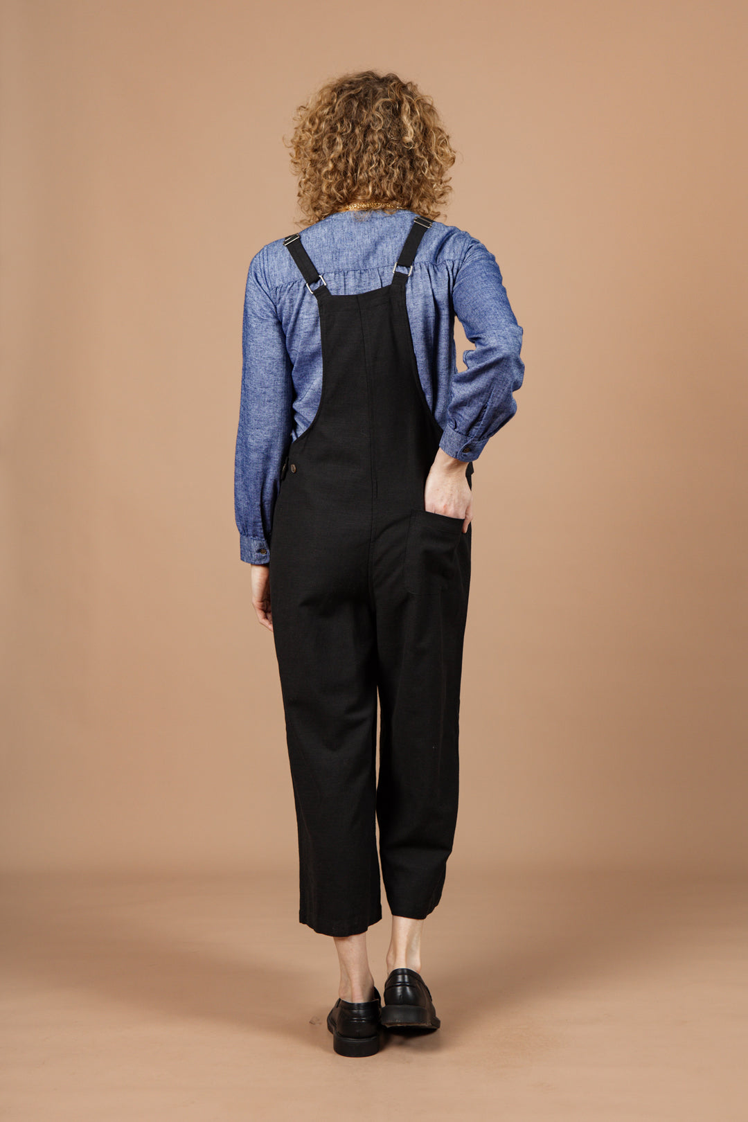 A person with curly hair stands facing away, wearing a blue long-sleeve shirt and the Bridge & Burn Edie Overall in Black with adjustable straps, paired with black shoes, against a plain beige background.