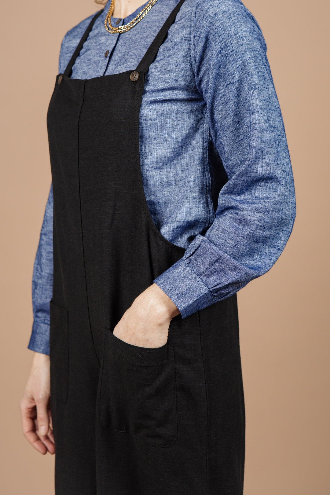 A person stands with one hand in their pocket against a plain beige background, wearing the Bridge & Burn Edie Overall in Black styled over a blue long-sleeve shirt.