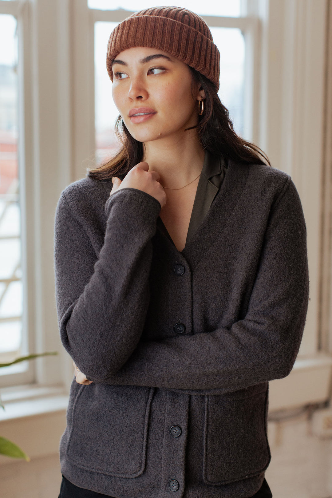 A woman wearing a brown knit beanie, gold hoop earrings, and the Bridge & Burn Whit Cardigan in Charcoal stands indoors by large windows, gazing thoughtfully to the side.