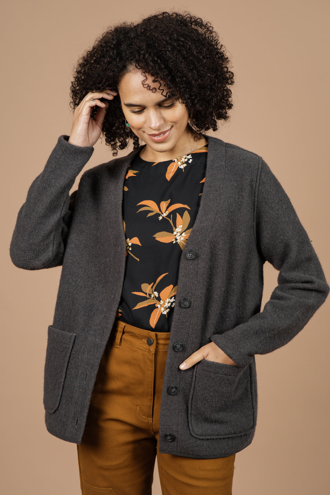A woman with curly hair, wearing the Bridge & Burn Whit Cardigan in Charcoal over a black shirt with an orange floral pattern and brown pants, stands smiling against a beige background and touches her hair.