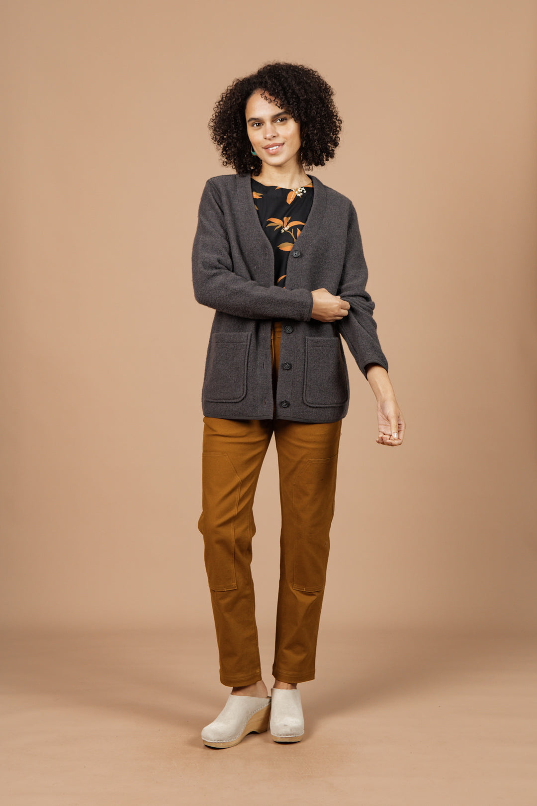 A woman with curly hair smiles against a beige background, wearing the Bridge & Burn Whit Cardigan in Charcoal over a patterned black top, mustard pants, and light shoes. She holds one arm with her other hand.