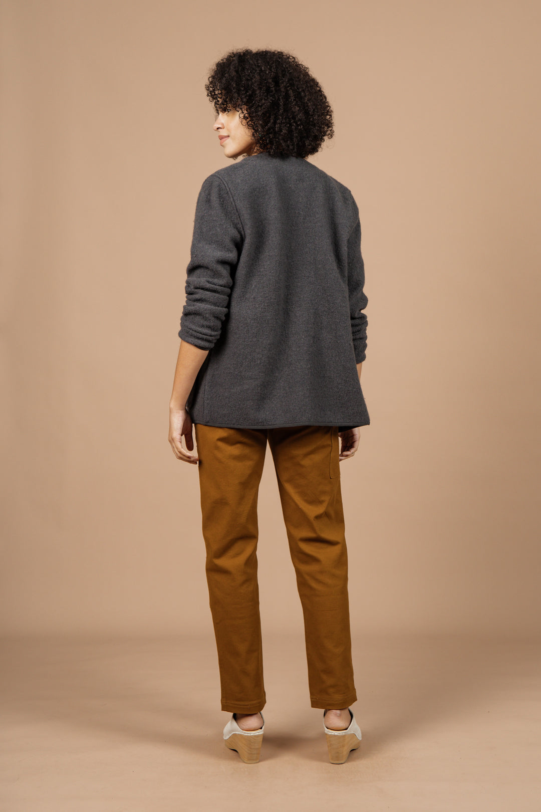 A person with curly hair, dressed in the Bridge & Burn Whit Cardigan in Charcoal, mustard pants, and beige shoes stands facing away against a solid beige background.