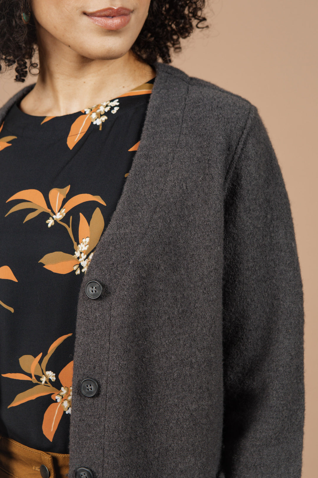 A woman wears the Bridge & Burn Whit Cardigan in Charcoal over a black, orange-floral shirt. Set against a beige background, only her lower face and torso are visible.