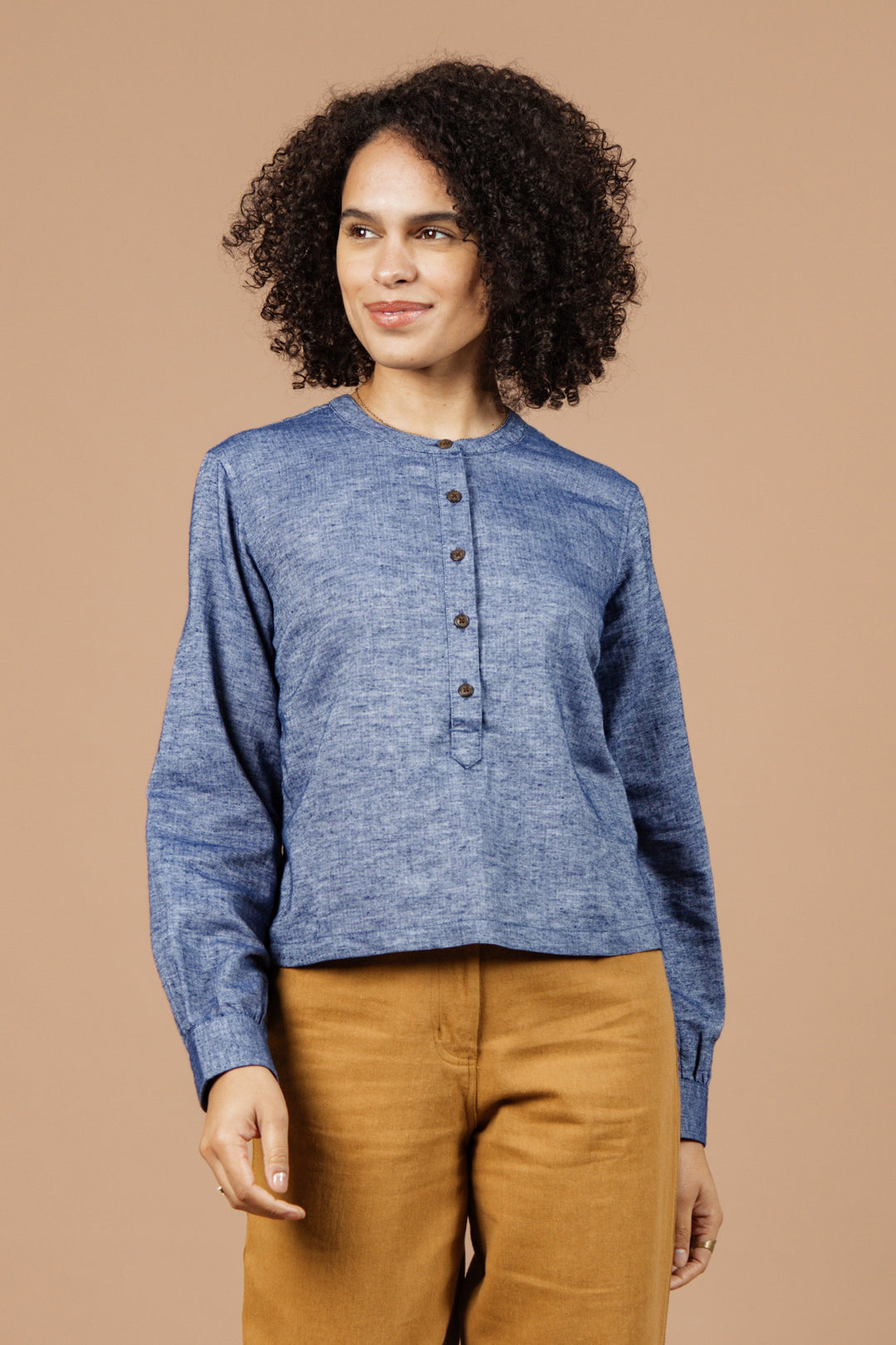 Wearing the Bridge & Burn Yarra Pullover in Indigo Herringbone, a person with curly hair and high-waisted tan pants stands against a beige background, glancing to the side with a slight smile—epitomizing sustainable style.