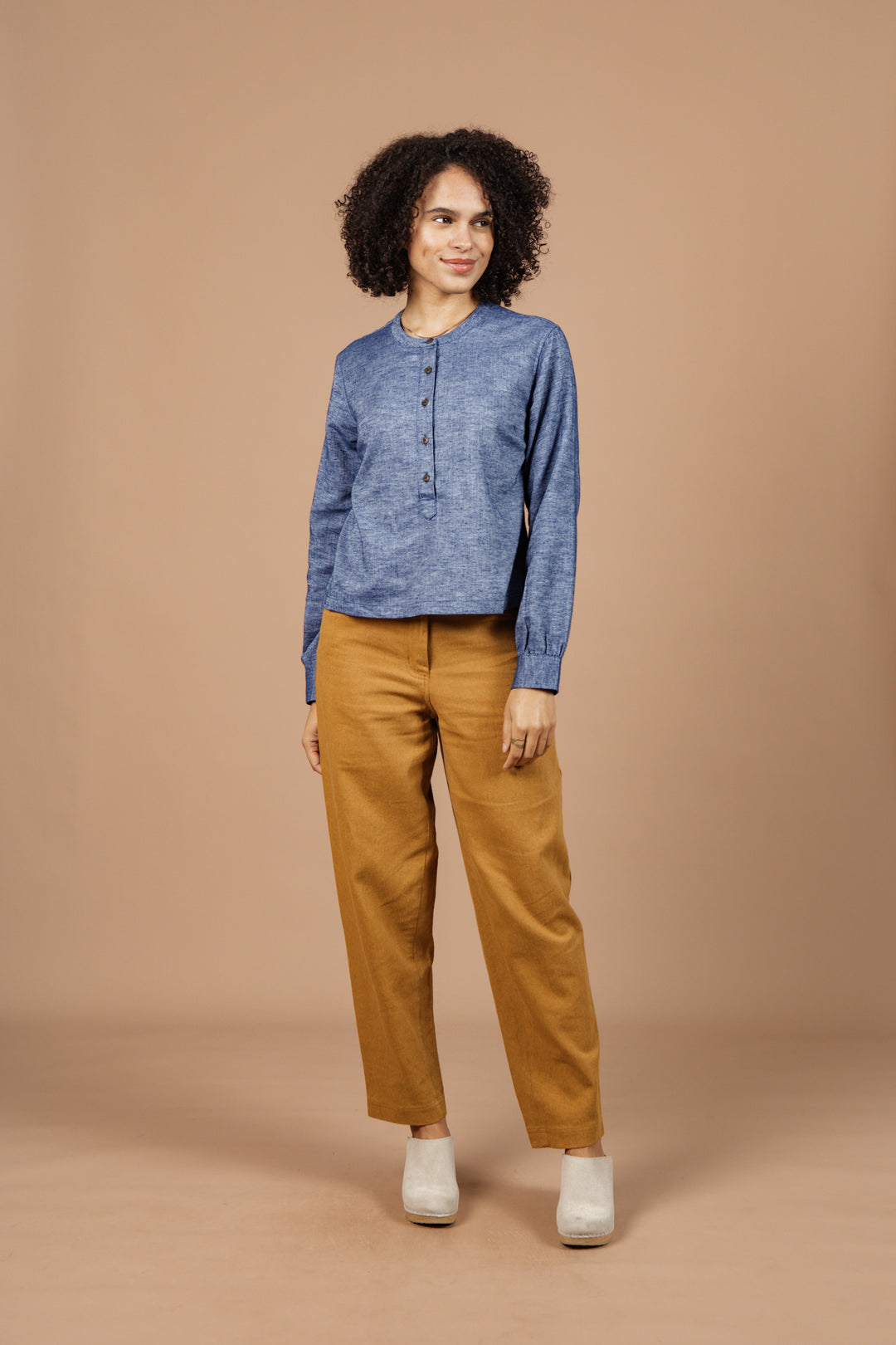 A person with curly hair stands against a beige background, smiling slightly and looking to the side, dressed in the Bridge & Burn Yarra Pullover in Indigo Herringbone—showcasing stylish, sustainable fashion.