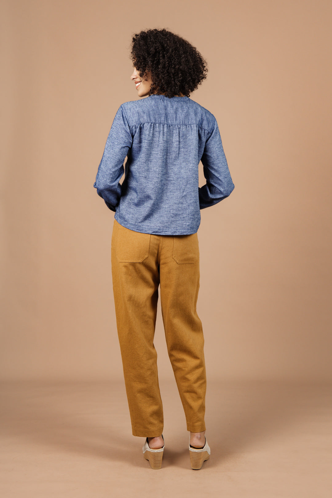 A person with curly hair, seen from behind, models the Bridge & Burn Yarra Pullover in Indigo Herringbone—crafted from organic cotton—paired with mustard pants and beige shoes, embodying sustainable style against a beige backdrop.