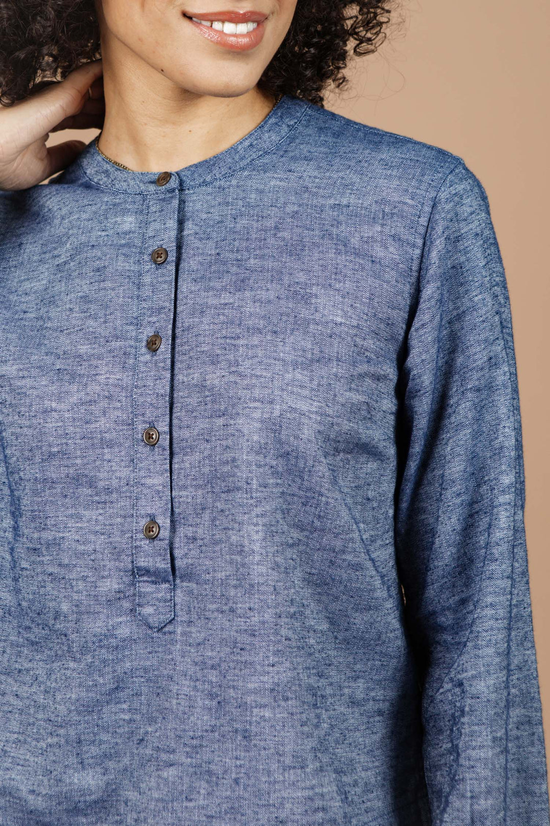 A person with curly hair wears the Bridge & Burn Yarra Pullover in Indigo Herringbone, smiling with one hand raised near their face against a neutral background. The photo shows their upper body and part of their face.