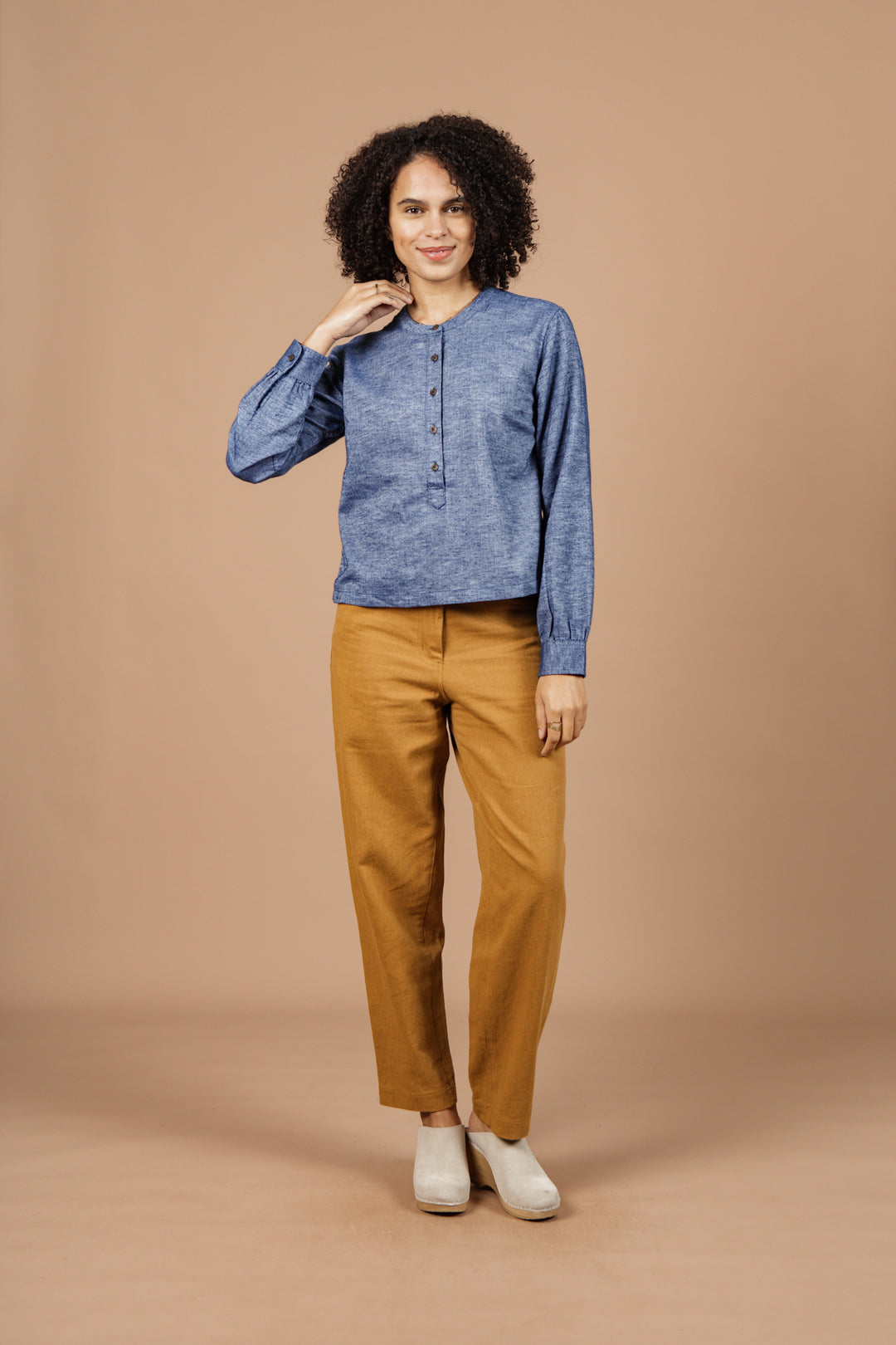 A person with curly hair wears the Bridge & Burn Yarra Pullover in Indigo Herringbone, paired with mustard yellow pants and white shoes, standing against a beige background and smiling while holding their collar.
