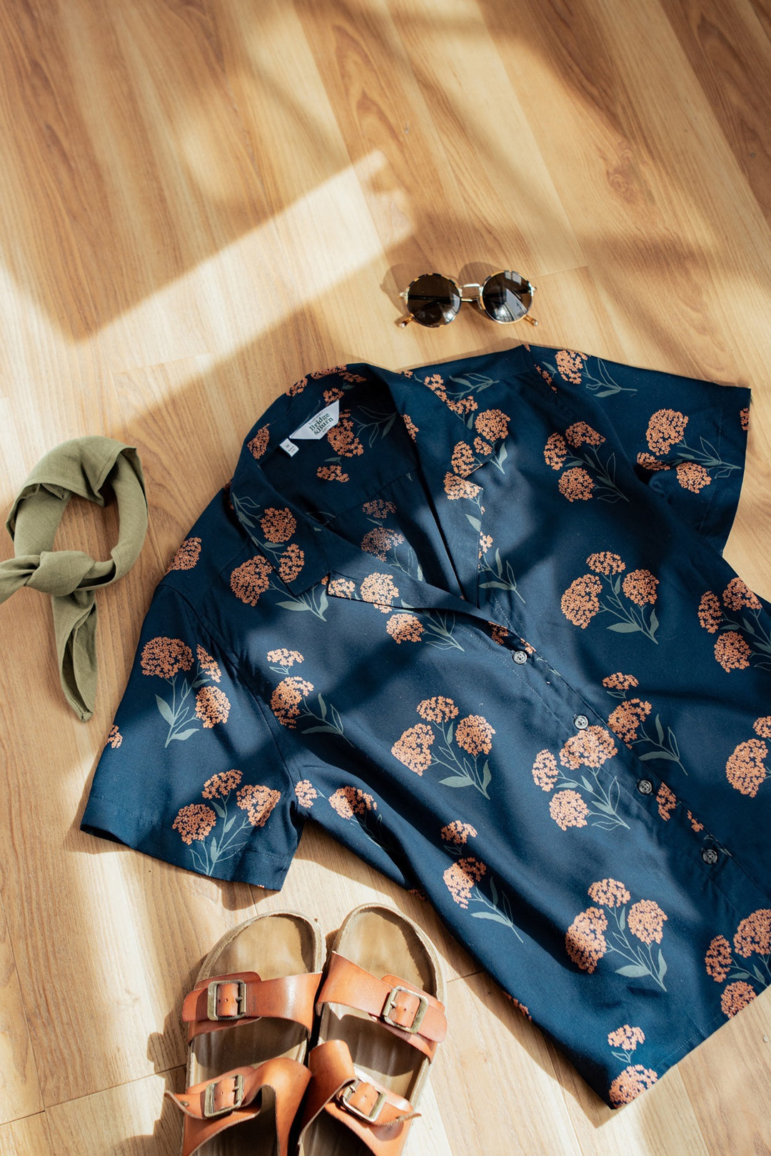 A flat lay of the Bridge & Burn Mila Cropped Shirt in Wildflower, tan sandals, a green hair tie, and round sunglasses arranged on a wooden floor with sunlight casting shadows.