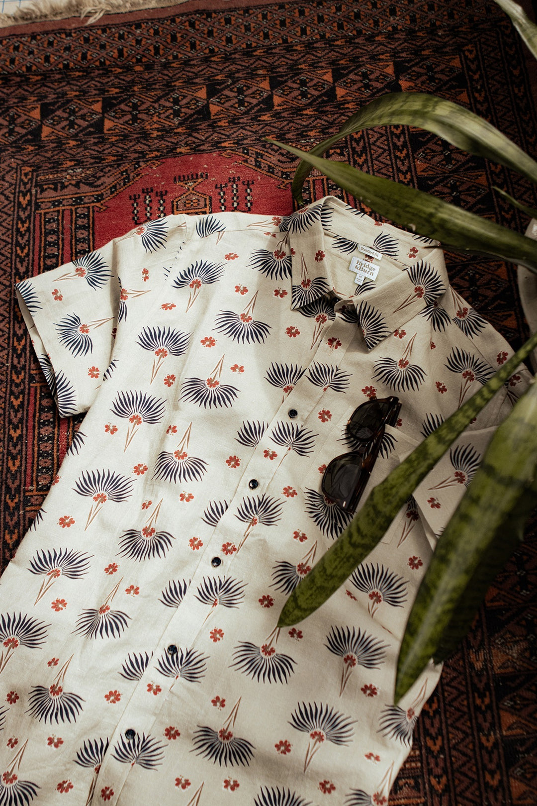 The Bridge & Burn Bea Shirt / Aloha Floral, a classic short-sleeve button down with white, blue, and orange floral print, is styled on a patterned rug with dark sunglasses and a green plant leaf in the foreground.