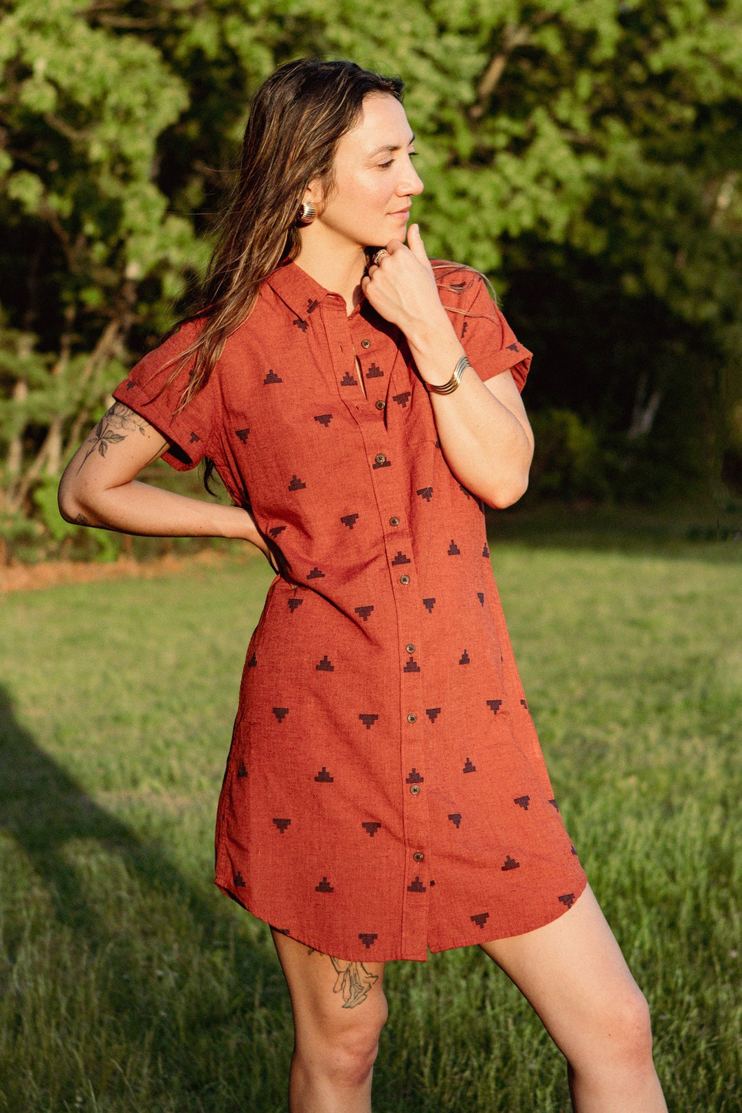 A woman stands on grass wearing the Bridge & Burn Loren Shirt Dress in Rust Fil-Coupé Pyramids—an organic cotton button-front dress with a subtle print. She has long brown hair, a bracelet, visible tattoos, and gazes thoughtfully to the side.