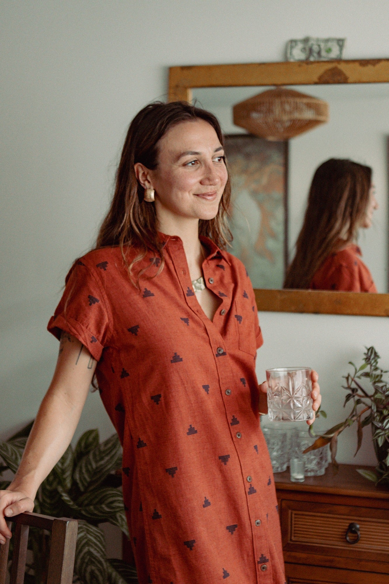 A woman in the Bridge & Burn Loren Shirt Dress in Rust Fil-Coupé Pyramids stands indoors, holding a glass of water and smiling softly, one hand on a chair. A mirror, plants, and wooden furniture appear in the background.
