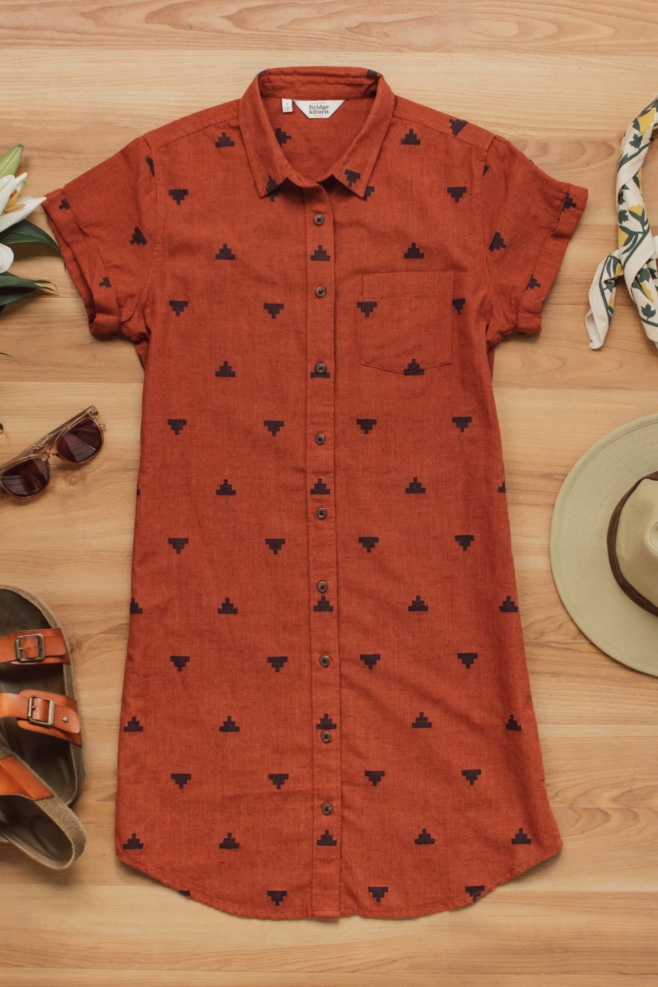 The Bridge & Burn Loren Shirt Dress in Rust Fil-Coupé Pyramids, an organic cotton button-front dress with a black geometric print, is displayed flat surrounded by sandals, sunglasses, flowers, a hat, and a patterned scarf.