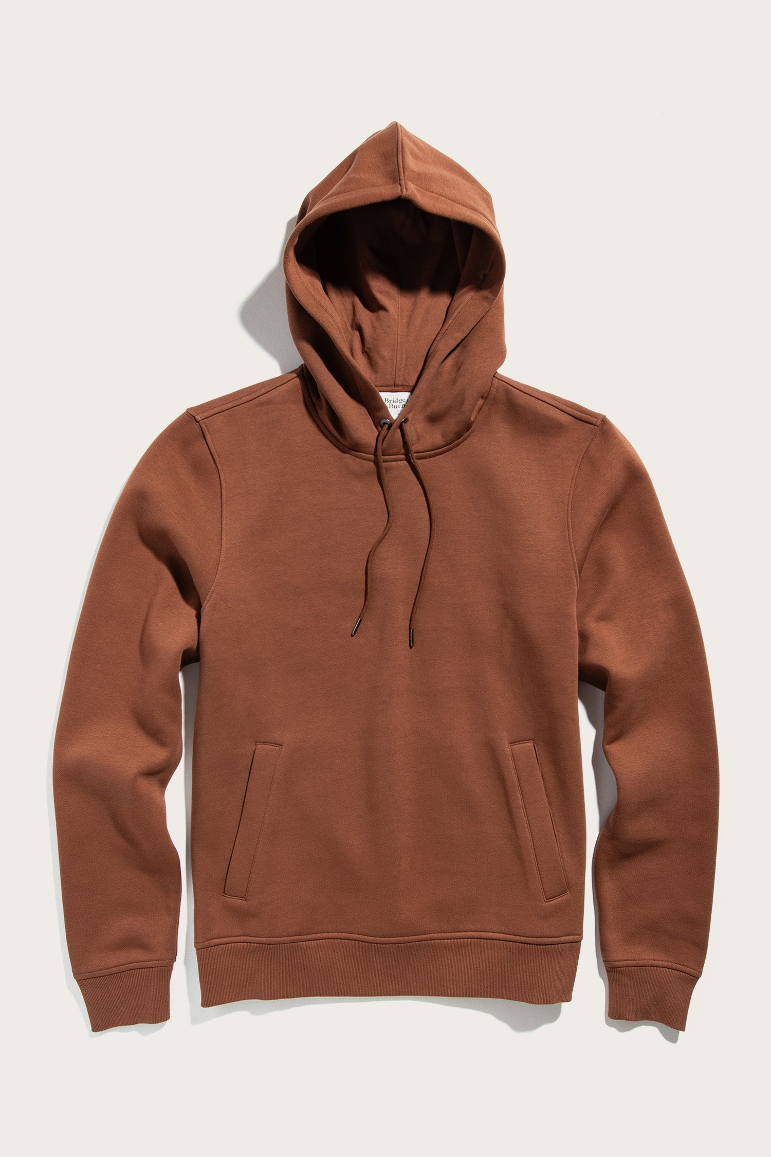 The Williams Pullover Hoodie in Burnt Umber by Bridge & Burn, made from organic cotton, features a front pouch pocket and drawstrings, shown laid flat on a light background.