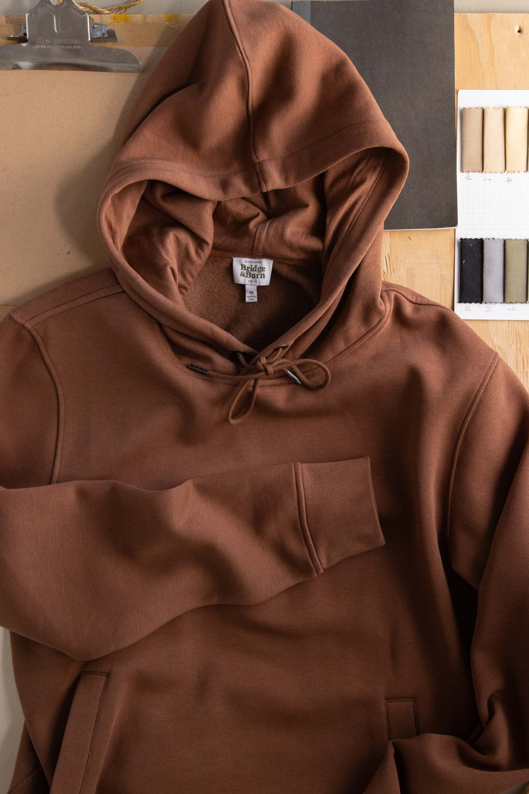 The Williams Pullover Hoodie in Burnt Umber by Bridge & Burn rests on a wooden surface beside fabric swatches, a clipboard, and paper. This organic cotton sweatshirt features a front pocket and hood with drawstring for eco-friendly comfort.