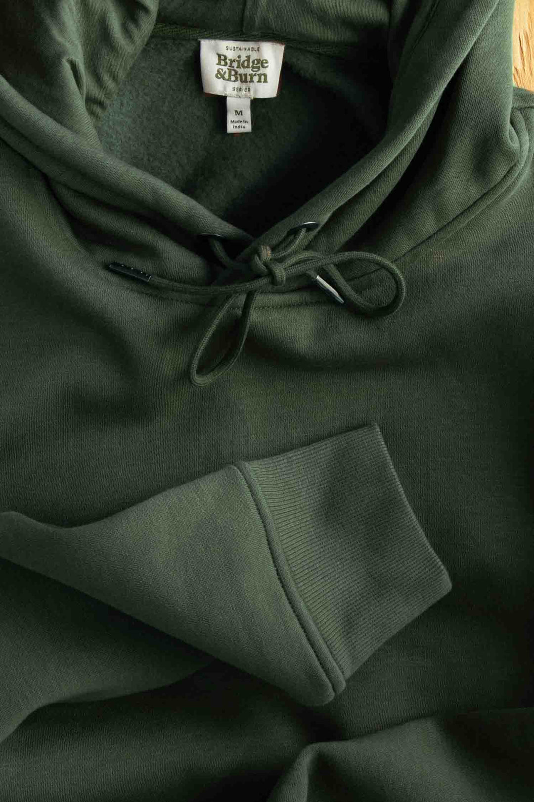 A close-up of the Bridge & Burn Williams Pullover Hoodie in Forest Night, made from recycled polyester, featuring drawstrings, a kangaroo pocket, and a visible label inside the collar.
