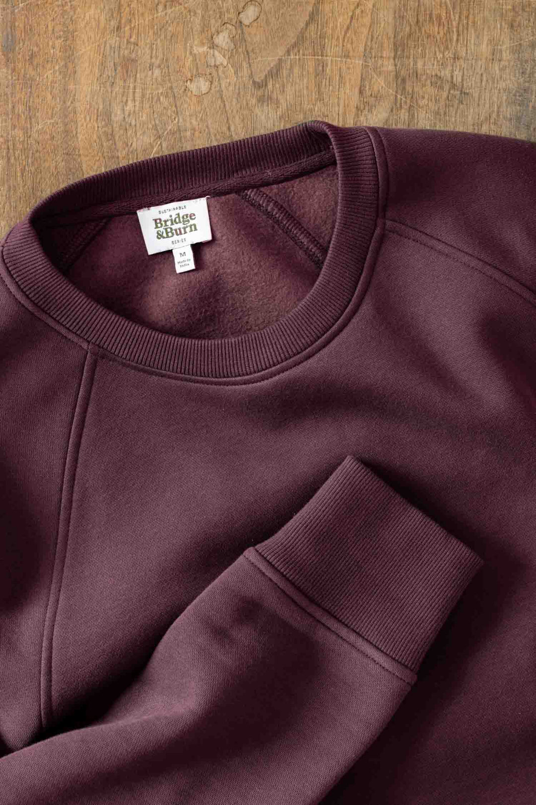 The Lincoln Crew Sweatshirt in Burgundy by Bridge & Burn, featuring a ribbed collar and cuffs plus brand label, is shown laid flat on a wooden surface.