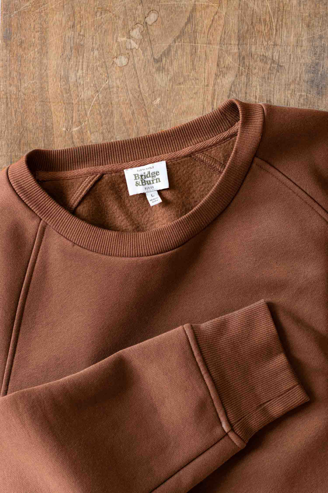 The Lincoln Crew Sweatshirt in Burnt Umber by Bridge & Burn is laid flat on wood, displaying its collar, sleeve, and a white Bridge & Burn tag.