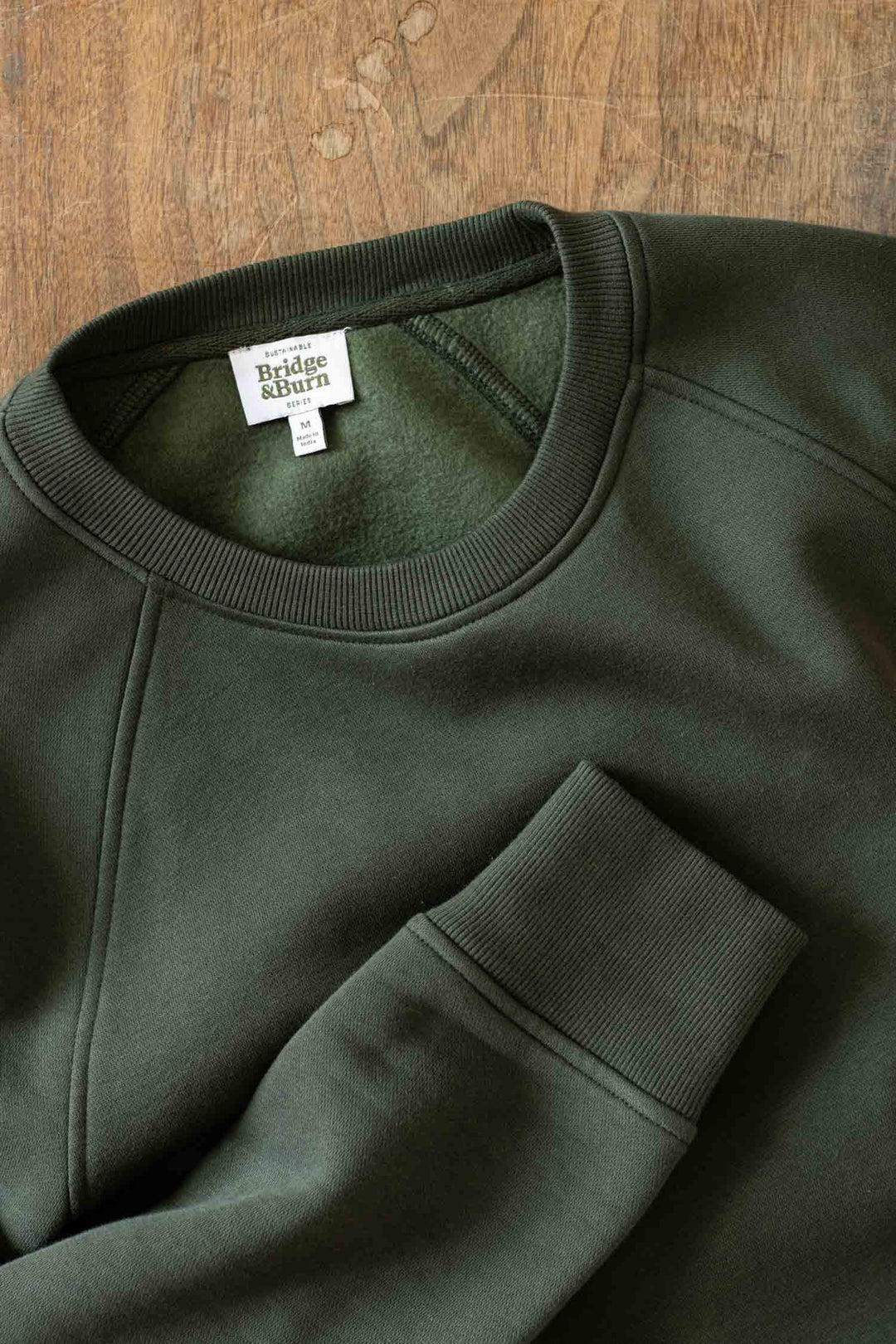 The Bridge & Burn Lincoln Crew Sweatshirt in Forest Night, featuring ribbed collar and cuffs along with a white brand label, is laid flat on a wooden surface.