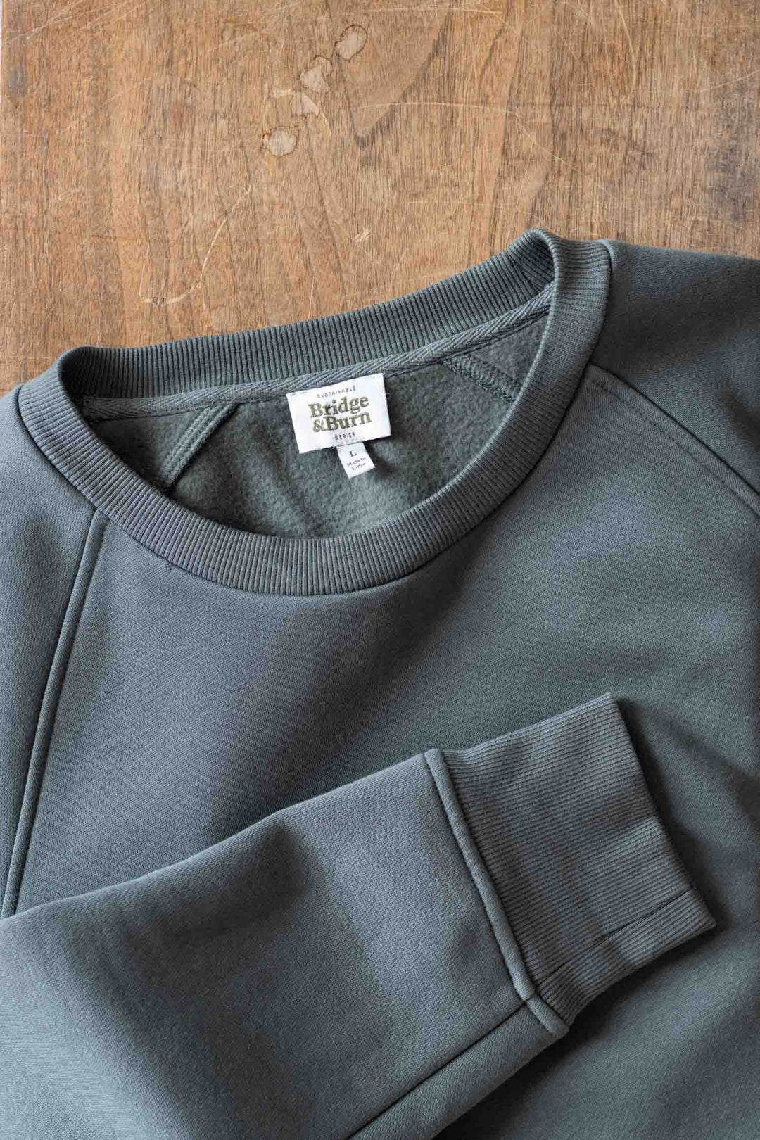 The Bridge & Burn Lincoln Crew Sweatshirt in Slate, a dark green sustainable crewneck with ribbed collar and cuffs, is displayed on a wooden surface with the sleeve folded across the body. The inside collar tag shows the brand name.