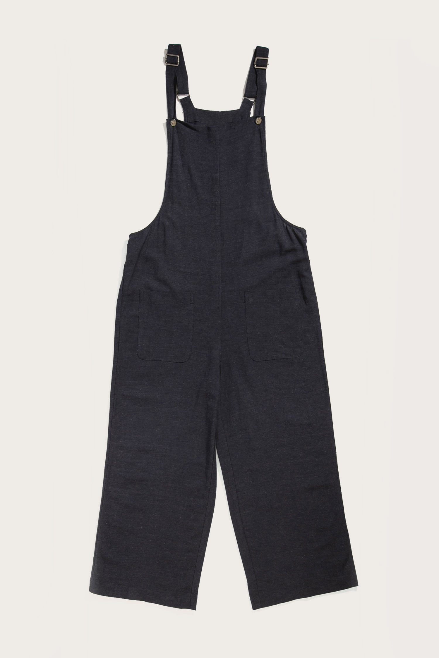 The Edie Overall in navy by Bridge & Burn is crafted from a soft linen-cotton blend, featuring adjustable shoulder straps and two large front pockets, displayed flat on a white background.