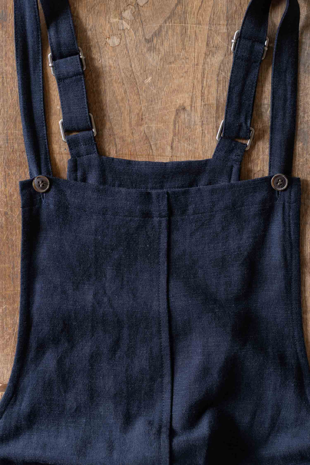 A close-up of the Bridge & Burn Edie Overall in navy, featuring wide legs, adjustable straps, and buttons, crafted from a soft linen-cotton blend, laid flat on a wooden surface.