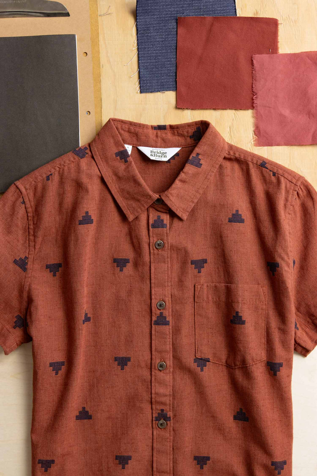 The Bridge & Burn Lana Shirt in Rust Fil-Coupé Pyramids, a sustainable short sleeve with a navy geometric pattern, is displayed on wood beside fabric swatches in black, dark blue, and two reds.