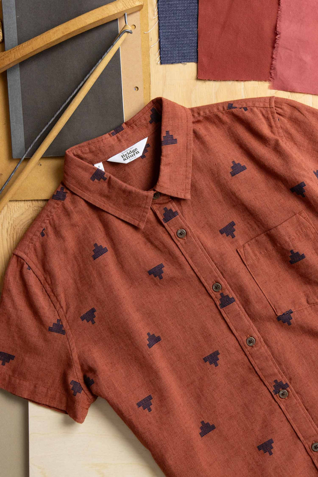 The Bridge & Burn Lana Shirt in Rust Fil-Coupé Pyramids, a sustainable short sleeve shirt made from organic cotton with navy jacquard geometric patterns, is displayed on wood beside fabric swatches and notebooks.