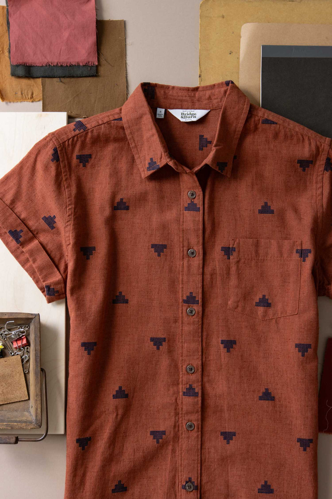 The Bridge & Burn Loren Shirt Dress in Rust Fil-Coupé Pyramids—a short-sleeve, button-front shirt dress with a small collar, chest pocket, and navy geometric motifs—laid flat amid fabric swatches and stationery.