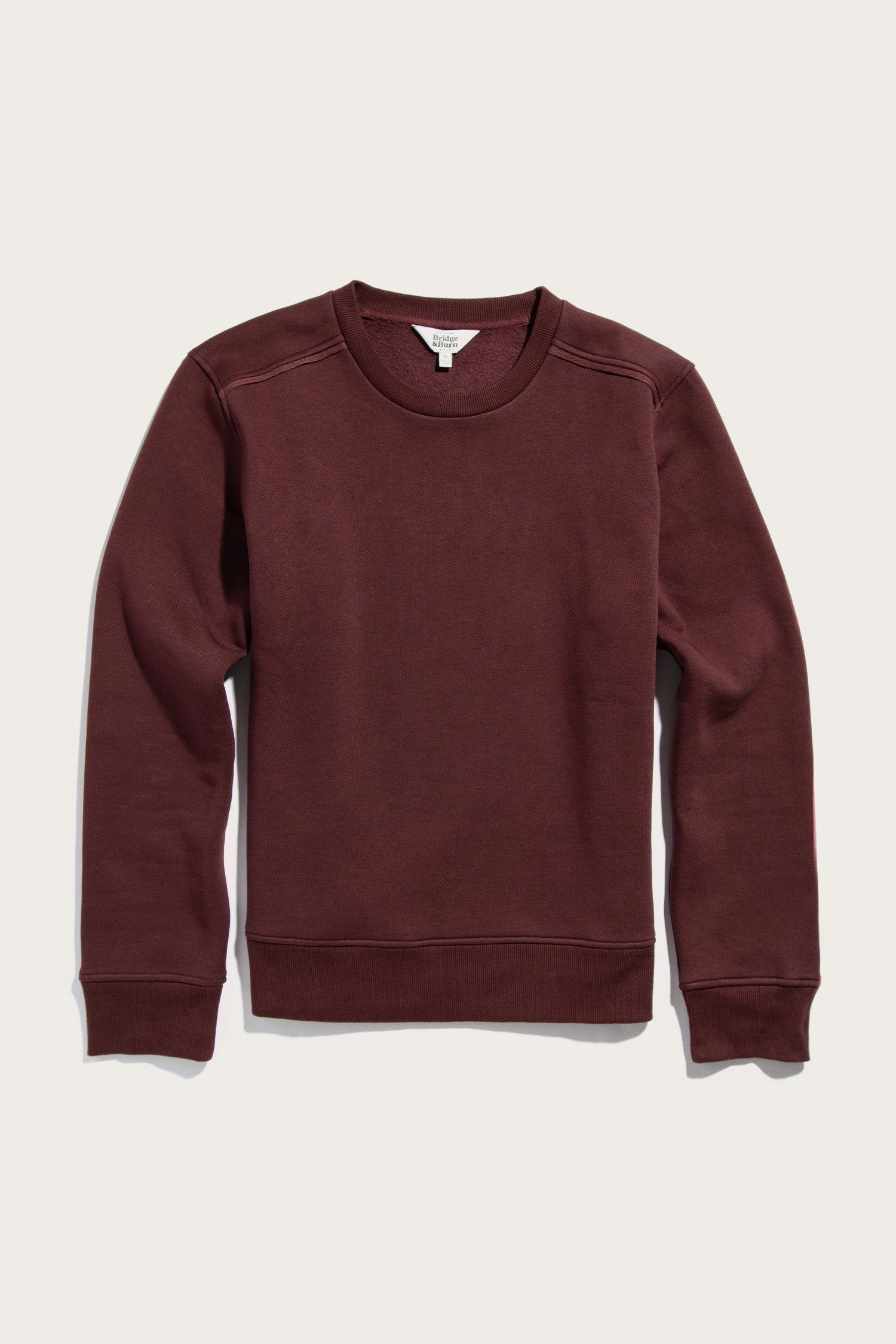 The Bailey Crew Sweatshirt by Bridge & Burn is a sustainable burgundy crewneck with long sleeves, ribbed cuffs, and a ribbed hem, shown laid flat on a light background.