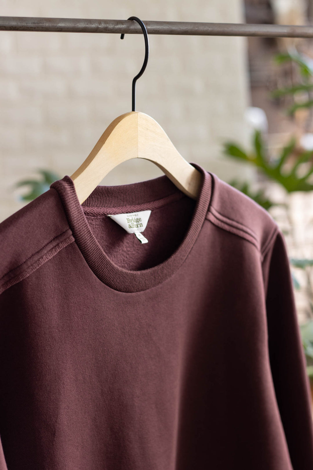 The Bailey Crew Sweatshirt in Burgundy by Bridge & Burn hangs on a wooden hanger from a metal rod, with greenery and a light wall blurred in the background.