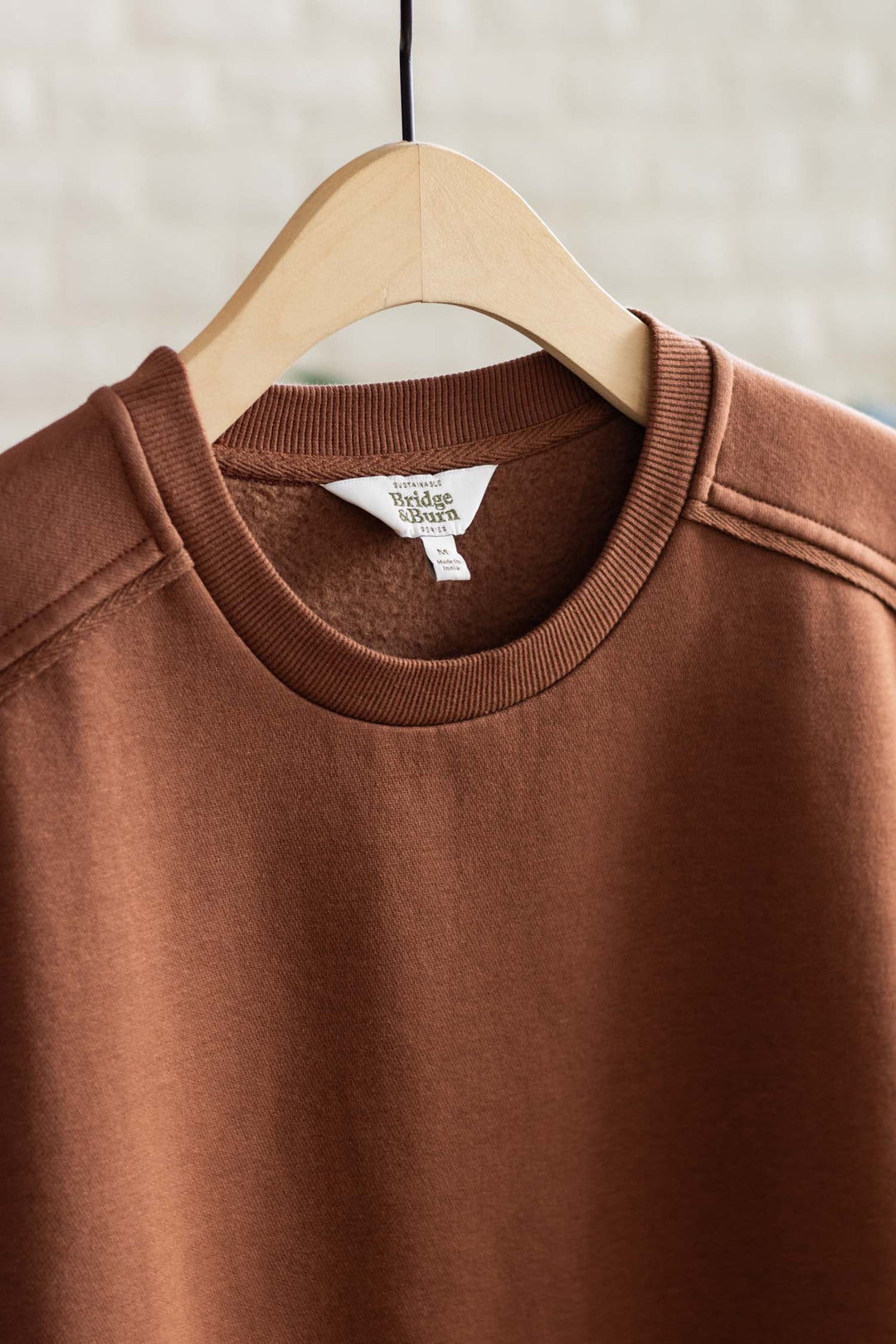 The Bailey Crew Sweatshirt in Burnt Umber by Bridge & Burn hangs on a wooden hanger, highlighting the neckline, shoulder details, and cozy fabric texture against a softly blurred light background.