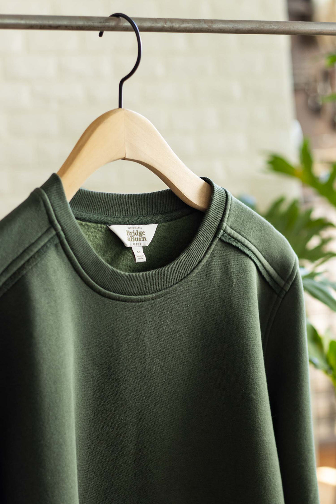 A close-up of the Bailey Crew Sweatshirt in Forest Night by Bridge & Burn, made from organic cotton, hanging on a wooden hanger with a blurred indoor background and green plants visible.